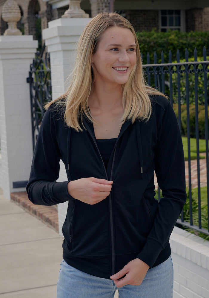 A young woman smiles outdoors in front of a white brick wall and black metal fence, wearing the Woolx Ryann Hoodie in black over a black top and light blue jeans. Her straight blonde hair is loose.