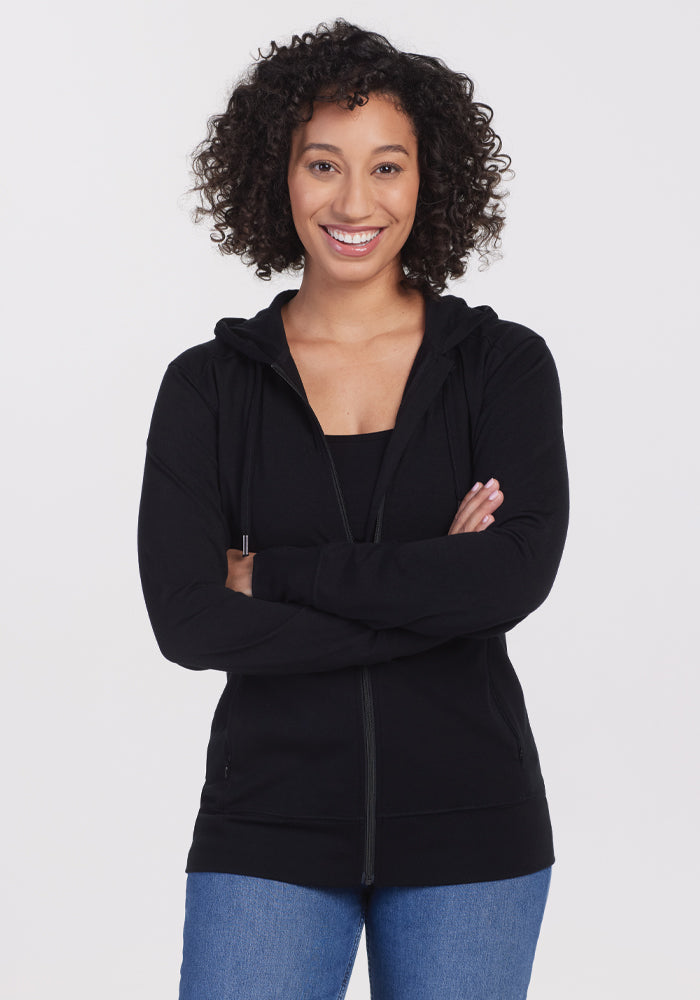 A person with curly hair stands against a white background, arms crossed casually, wearing a Woolx Ryann Hoodie in black paired with blue jeans, perfectly blending comfort and style. | Tori is 5'7", wearing a size S