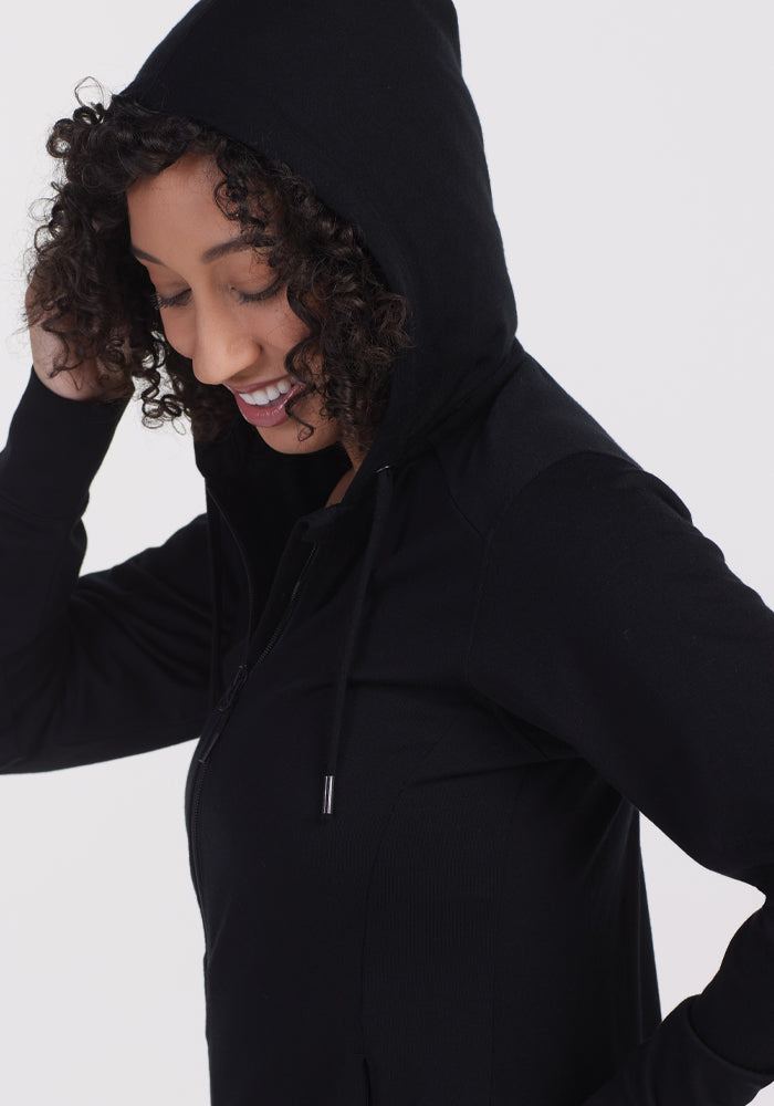 A person with curly hair smiles while wearing the Woolx Ryann Hoodie in black, which lightly covers part of their head. They stand against a plain, light gray background.