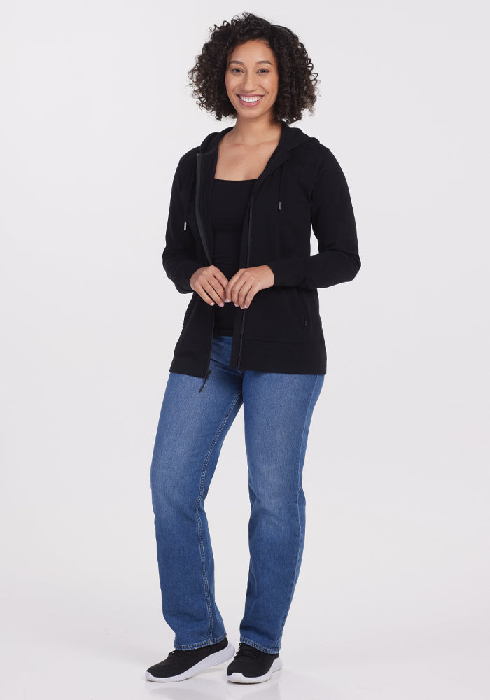 A person with short curly hair smiles, wearing the Woolx Ryann Hoodie in black over a black top, blue jeans, and black sneakers. They stand relaxed against a plain white background, showcasing their travel essential attire.