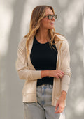A woman with long blonde hair and sunglasses smiles as she stands against a light wall, wearing the Woolx Ryann Hoodie in Cream Heather—her perfect travel essential—paired with a black t-shirt and light blue jeans.