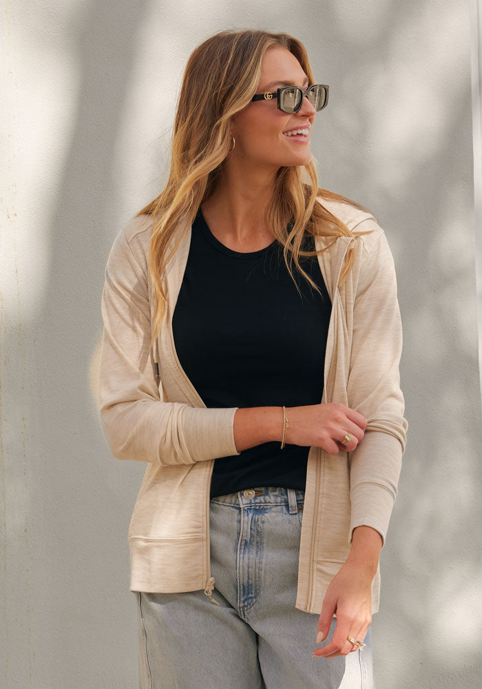 A woman with long blonde hair and sunglasses smiles as she stands against a light wall, wearing the Woolx Ryann Hoodie in Cream Heather—her perfect travel essential—paired with a black t-shirt and light blue jeans.