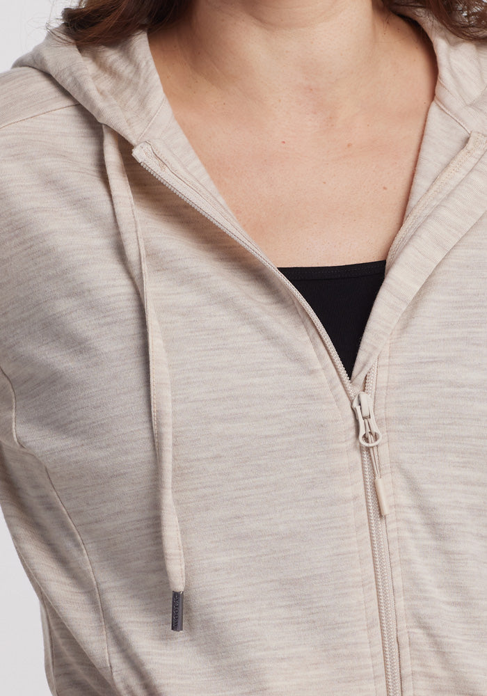 A close-up shows a person wearing the Woolx Ryann Hoodie in Cream Heather, crafted from lightweight merino wool with a drawstring and subtle heather pattern over a black top. The hoodie is perfect for travel, though the person's face isn’t visible.