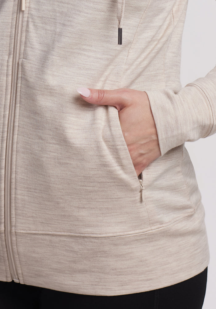 A close-up shows a person wearing the Woolx Ryann Hoodie in Cream Heather, crafted from soft merino wool with a subtle texture. Their hand rests in the front pocket, paired stylishly with black pants, and their manicured nails add to this travel essential's practicality.