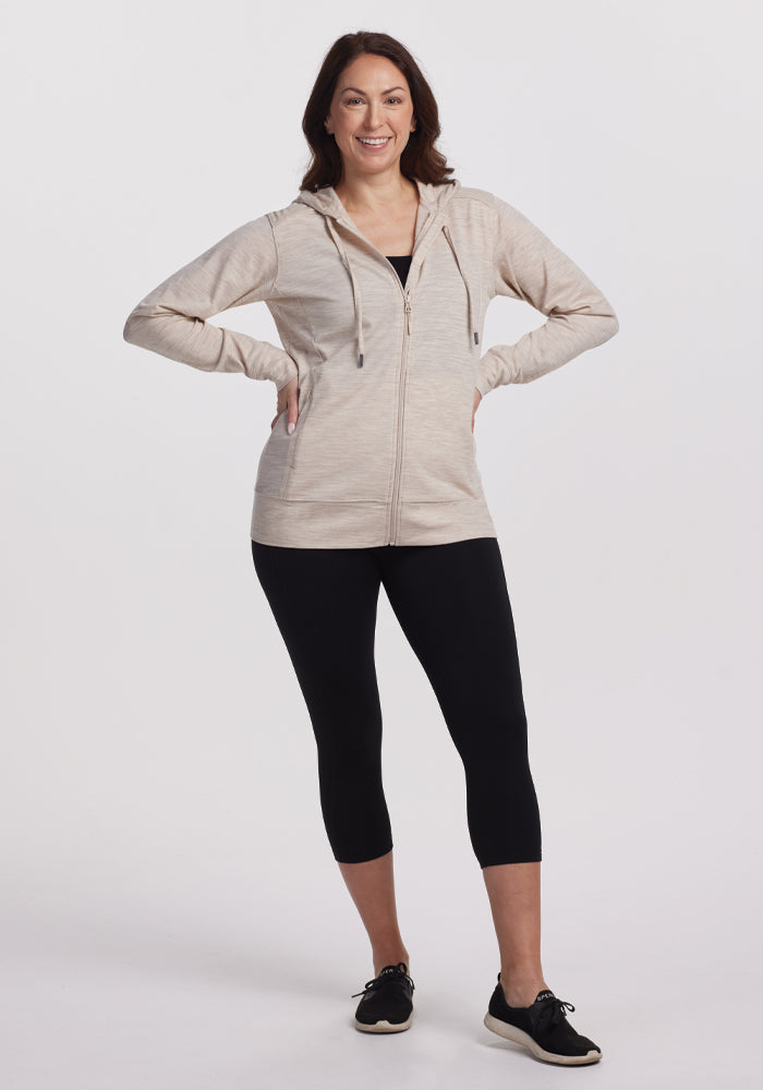 A person in Woolx's Ryann Hoodie in Cream Heather and black leggings stands smiling with hands on their hips. They wear moisture-wicking black shoes against a plain white background.
