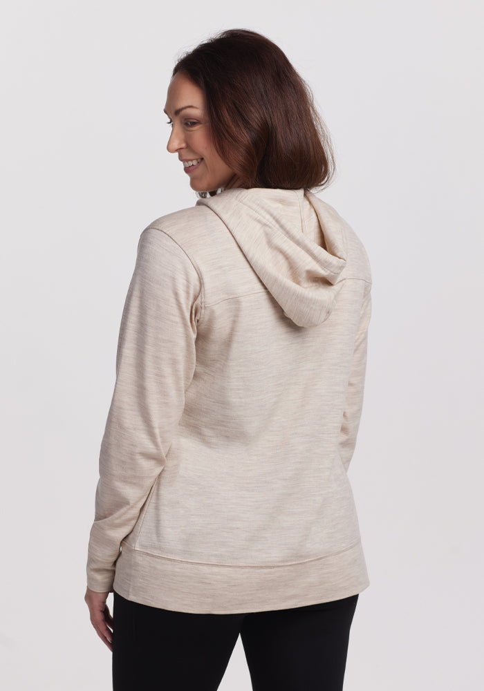 A woman models the Woolx Ryann Hoodie in Cream Heather, smiling over her shoulder. This lightweight travel essential has a cozy hood and simple design, ideal for adventures. She pairs it with black pants against a plain white background.
