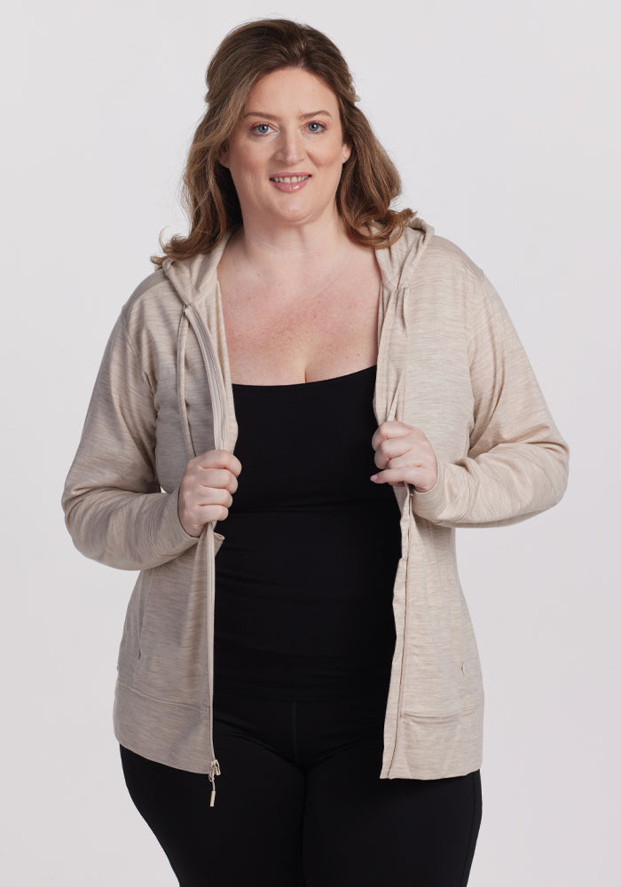 A smiling person with wavy hair stands against a plain background, wearing Woolx's Ryann Hoodie in Cream Heather over a black top and pants. They hold the hoodie open with both hands, highlighting its cozy yet breathable design. | Cambre is 5'11", wearing a size XL