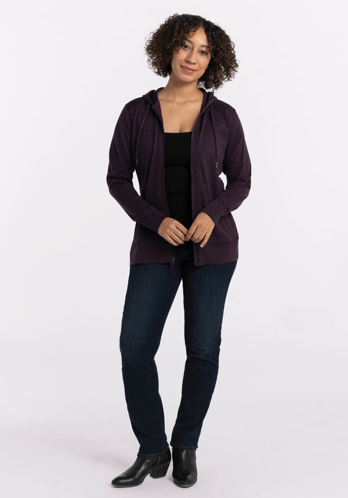 Person standing against a plain background, wearing a Woolx Ryann Hoodie in Deep Plum over a black top, blue jeans, and ankle boots. They have short, curly hair and are looking at the camera with hands resting in front. Final Sale Deals!.