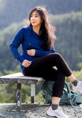 Woman in the Woolx Ryann Hoodie in Estate Blue sits outdoors with her backpack, surrounded by trees and mountains.