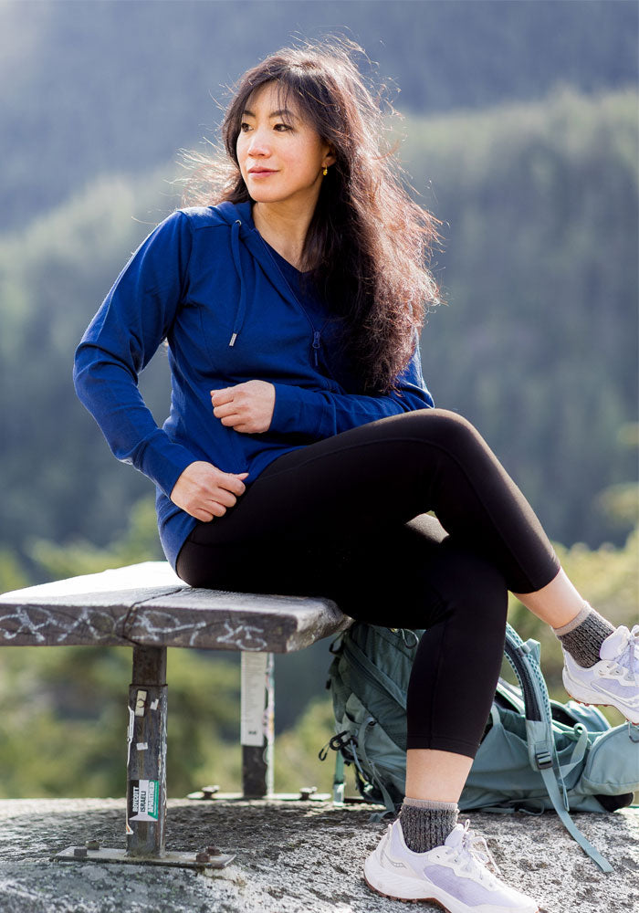 Woman in the Woolx Ryann Hoodie in Estate Blue sits outdoors with her backpack, surrounded by trees and mountains.