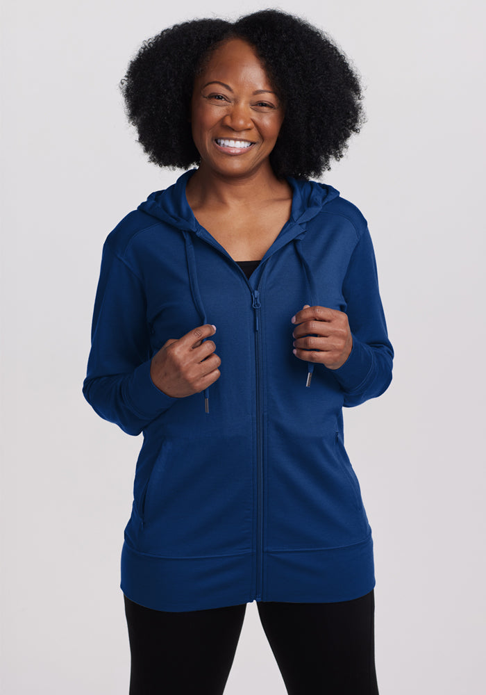 A smiling woman wears the Woolx Ryann Hoodie in Estate Blue and black pants against a plain light background.