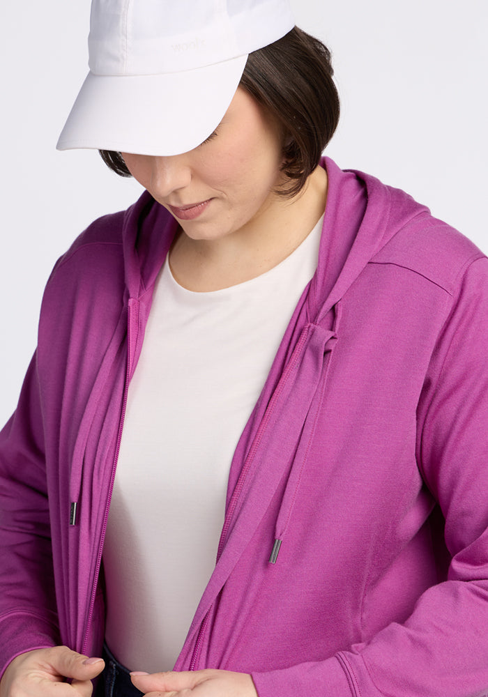 A woman in a white cap and shirt adjusts her Woolx Ryann Hoodie in Purple Wine, a comfy travel essential.