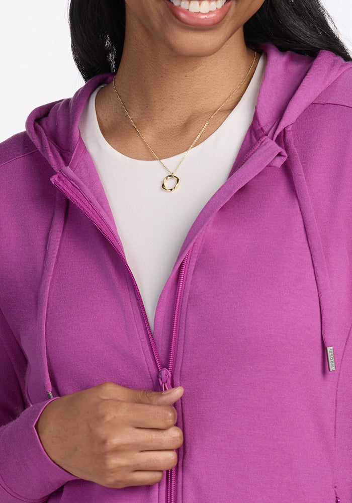 A woman smiles, holding the zipper of the purple Woolx Ryann Hoodie. She wears a white shirt and a gold necklace.