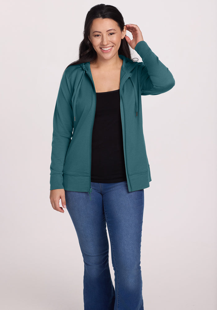 A woman in the Woolx Ryann Hoodie in Tidepool smiles, touching her hair against a plain background.