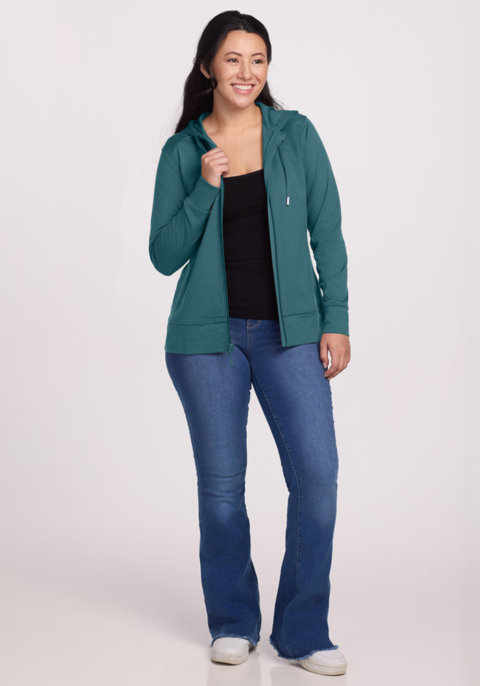 Woman smiling in a Woolx Ryann Hoodie in Tidepool, black top, blue jeans, and white sneakers.