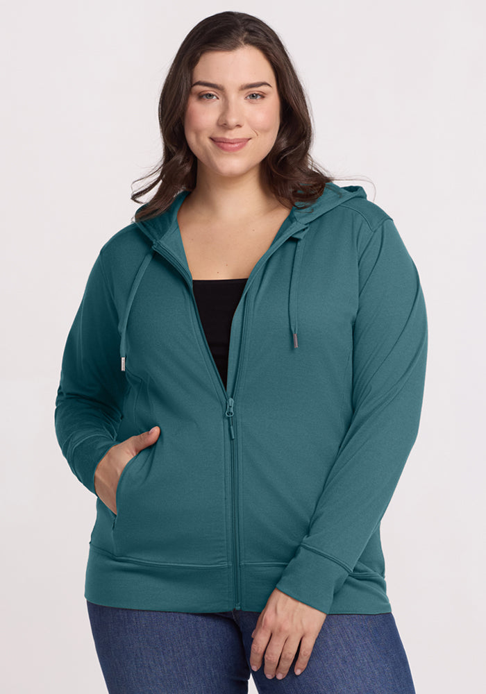 A woman smiles in the Woolx Ryann Hoodie • Final Sale - Tidepool and jeans, nailing a travel-essential look.