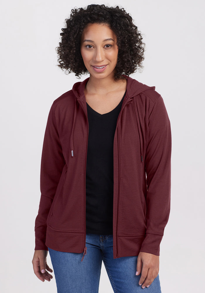 Woman with curly hair wearing a Woolx Ryann Hoodie in Toasted Garnet, black shirt, and blue jeans, smiling at the camera.