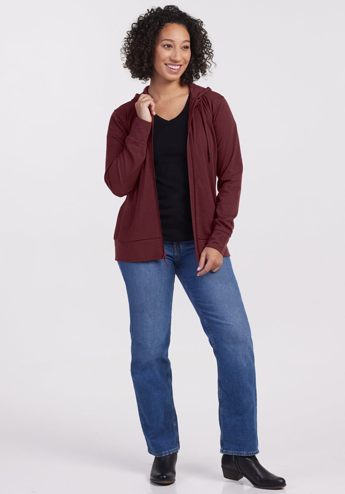 Smiling woman in a Woolx Ryann Hoodie in Toasted Garnet, black shirt, blue jeans, and boots against a white background.