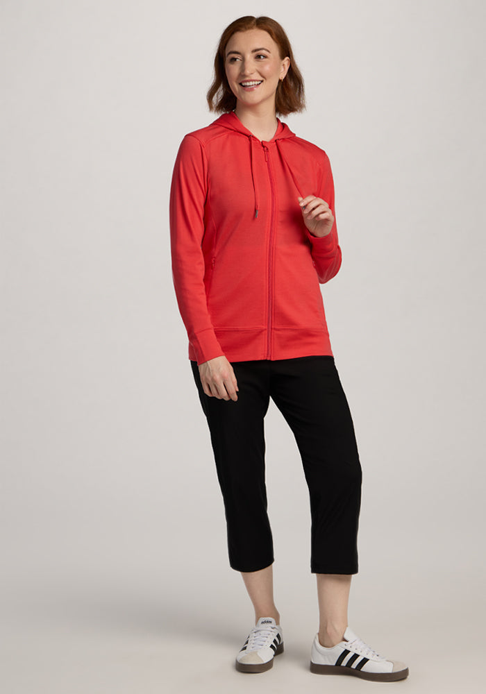 Woman wears the Woolx Ryann Hoodie in Hibiscus, paired with black cropped pants and white sneakers, smiling.