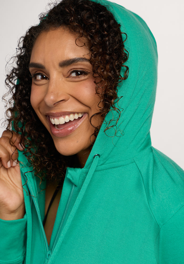 Smiling woman with curly hair wears the Woolx Ryann Hoodie - Jungle, holding the hood—a travel essential.