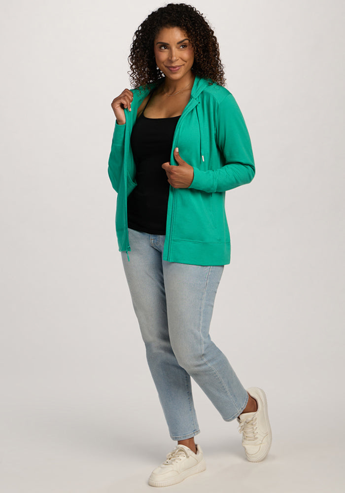 Woman smiling in the Woolx Ryann Hoodie - Jungle, black top, light blue jeans, and white sneakers.