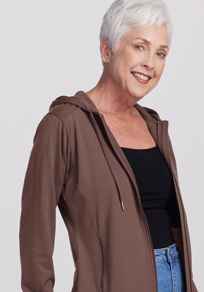 Smiling older woman in a Woolx Ryann Hoodie - Peppercorn, black top, and blue jeans against a light background.