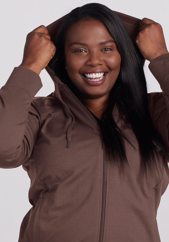 Smiling woman in a Woolx Ryann Hoodie - Peppercorn, a moisture-wicking travel essential, faces the camera.