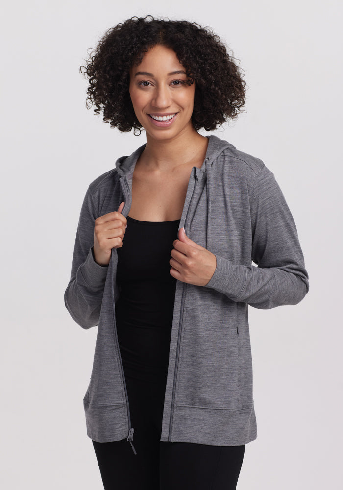 A person with curly hair wears the Woolx Ryann Hoodie - Graphite Heather over a black top, smiling against a white background. Known for its moisture-wicking properties, the lightweight travel hoodie is gripped by both hands. | Tori is 5'7", wearing a size S