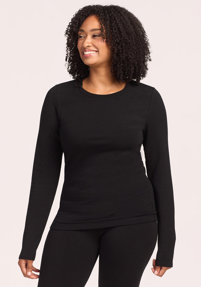 A woman with curly hair smiles while wearing the Woolx Sadie Baselayer Top in black and matching pants, standing against a light, neutral background. | Maysa is 5’3”, wearing a size M