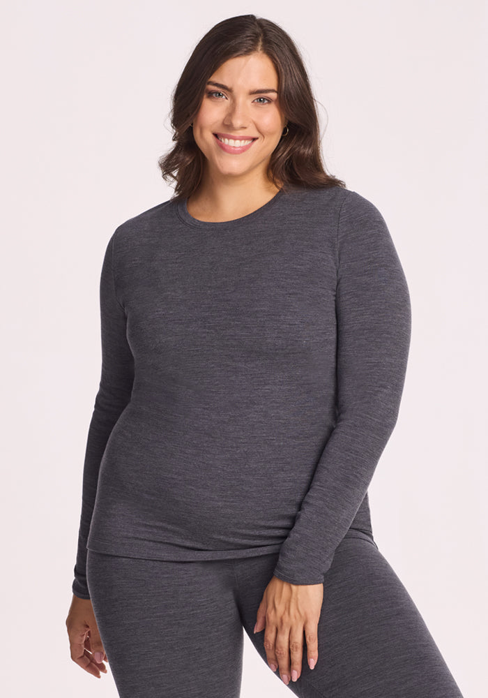 A woman with wavy brown hair smiles while wearing the Woolx Sadie Baselayer Top in Cinder Melange, paired with matching bottoms, standing against a plain light background. | Juliana is 5'10", wearing a size XL