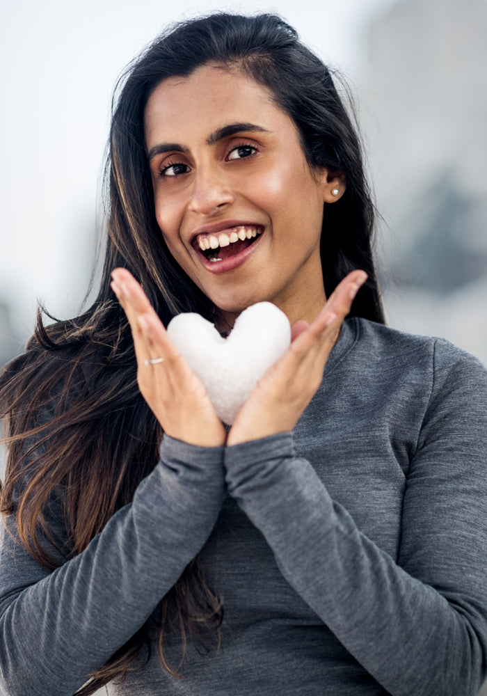 A smiling woman wears the Woolx Sadie Baselayer Top in gray as she holds a heart-shaped snowball outdoors, radiating warmth and comfort against a blurred background.