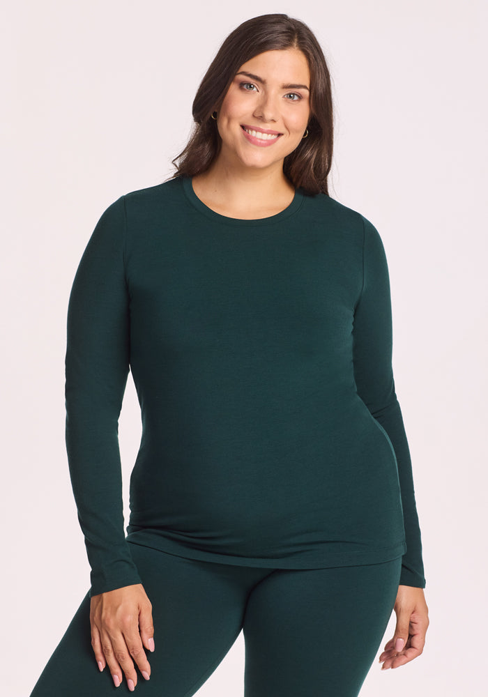 A woman with long brown hair, wearing the Woolx Sadie Baselayer Top in Deep Timber, stands and smiles against a plain light background.