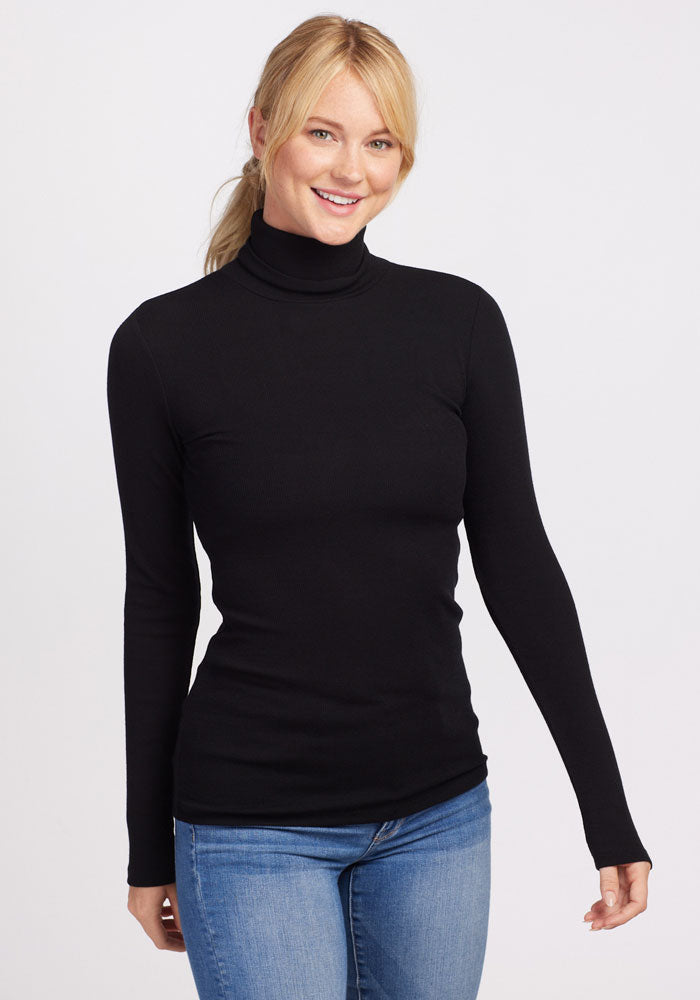 A person with long hair tied back is smiling, wearing a black Sage Ribbed Turtleneck from Woolx and blue jeans against a plain white background.