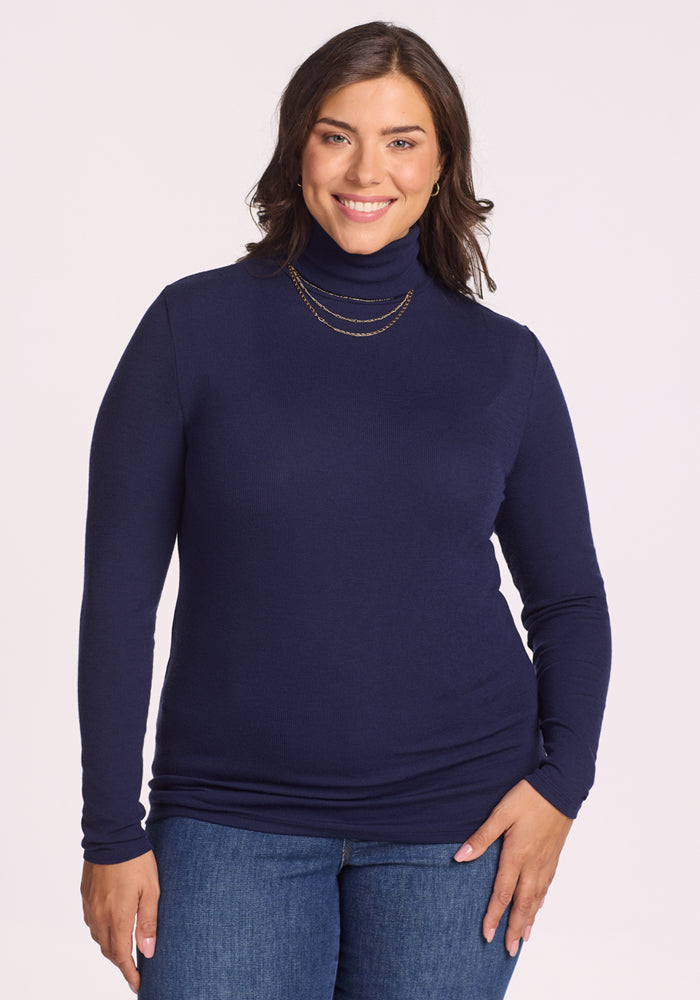 A woman with dark hair smiles and poses against a light background, wearing the Woolx Sage Ribbed Turtleneck in Midnight Sapphire and blue jeans, accessorized with a gold layered necklace.