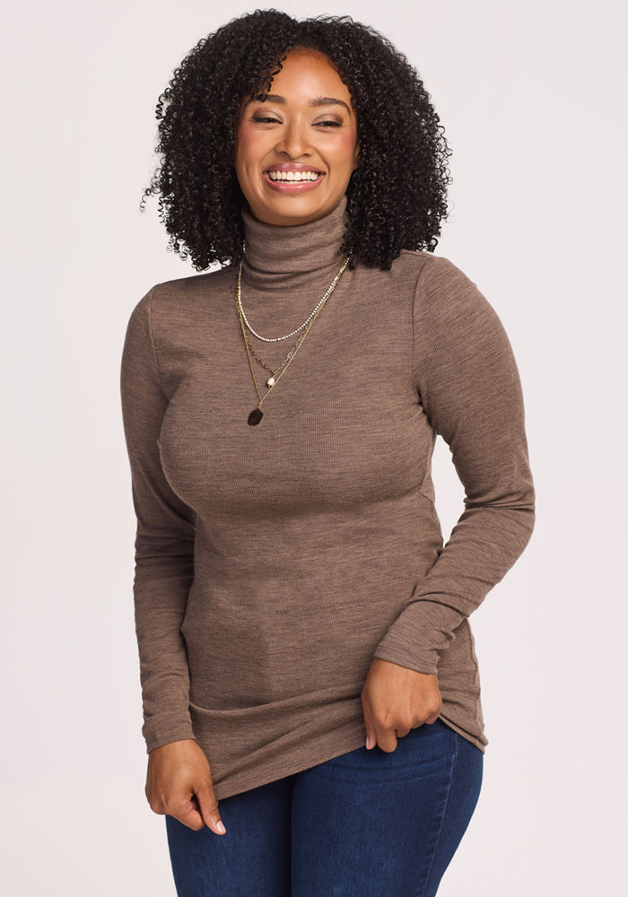 A woman with curly hair smiles while wearing the Woolx Sage Ribbed Turtleneck in Simply Taupe, layered gold necklaces, and blue jeans, standing against a plain light background.