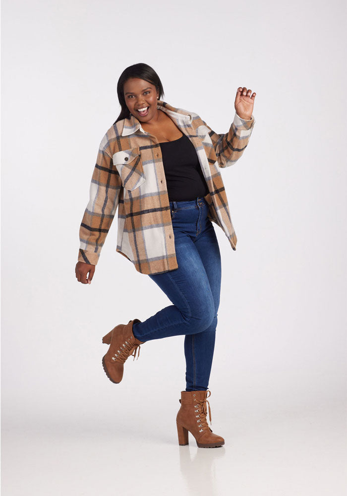 A woman models the Woolx Sawyer Shacket in Camel Plaid, paired with a black top, blue jeans, and brown heeled boots. She stands on one leg and smiles playfully against a plain white background.