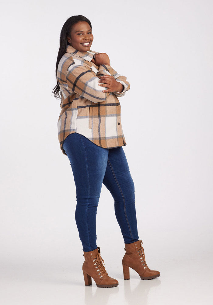 A woman smiles in the Woolx Sawyer Shacket - Camel Plaid, paired with blue skinny jeans and brown heeled lace-up boots, posing with one hand on her chin against a plain white background.
