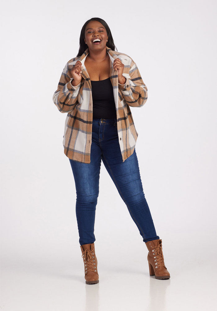 A smiling woman poses in the Woolx Sawyer Shacket - Camel Plaid, paired with a black top, blue jeans, and brown lace-up boots against a plain white background.