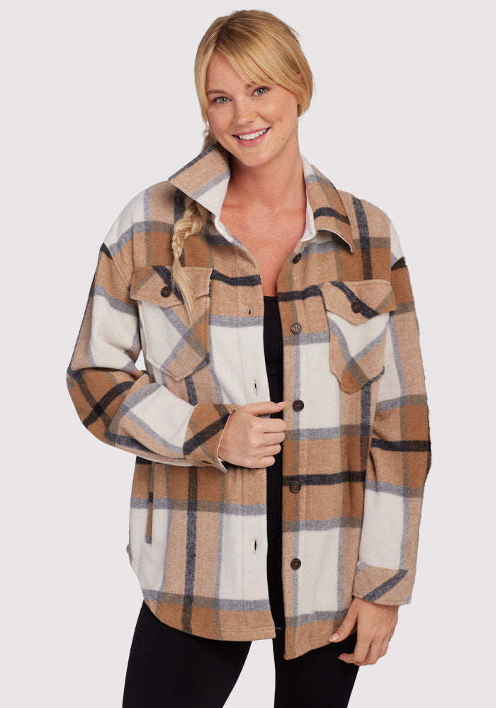 A smiling person with blonde hair in a side braid wears the Woolx Sawyer Shacket in Camel Plaid over a black top, standing against a plain light background.