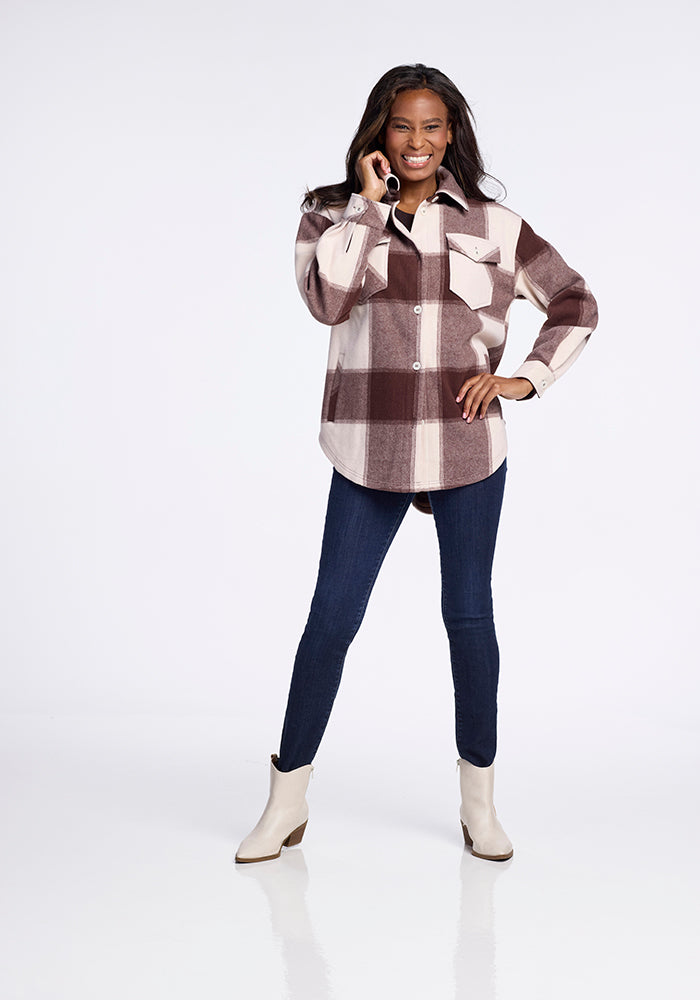 A woman smiles while talking on the phone, wearing the Woolx Sawyer Shacket in Chai Latte Plaid over dark skinny jeans and light ankle boots. The background is plain white.
