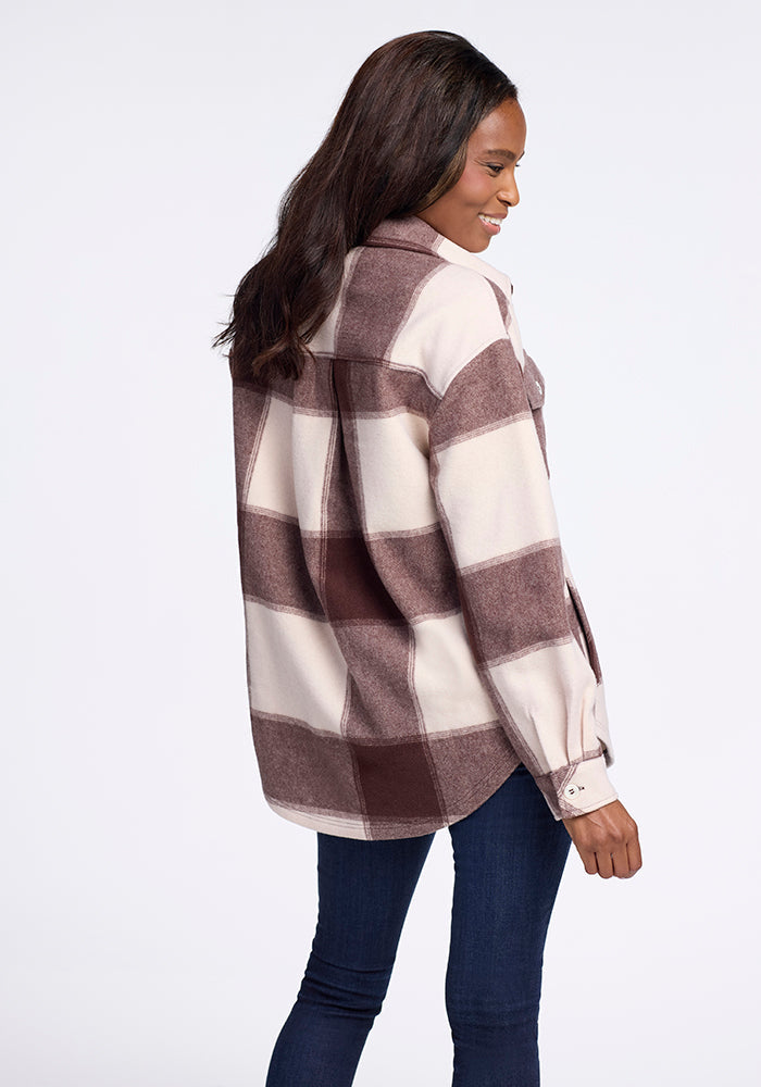 A woman with long dark hair, wearing the Woolx Sawyer Shacket in Chai Latte Plaid and dark blue jeans, stands turned to the side and smiles against a plain white background.