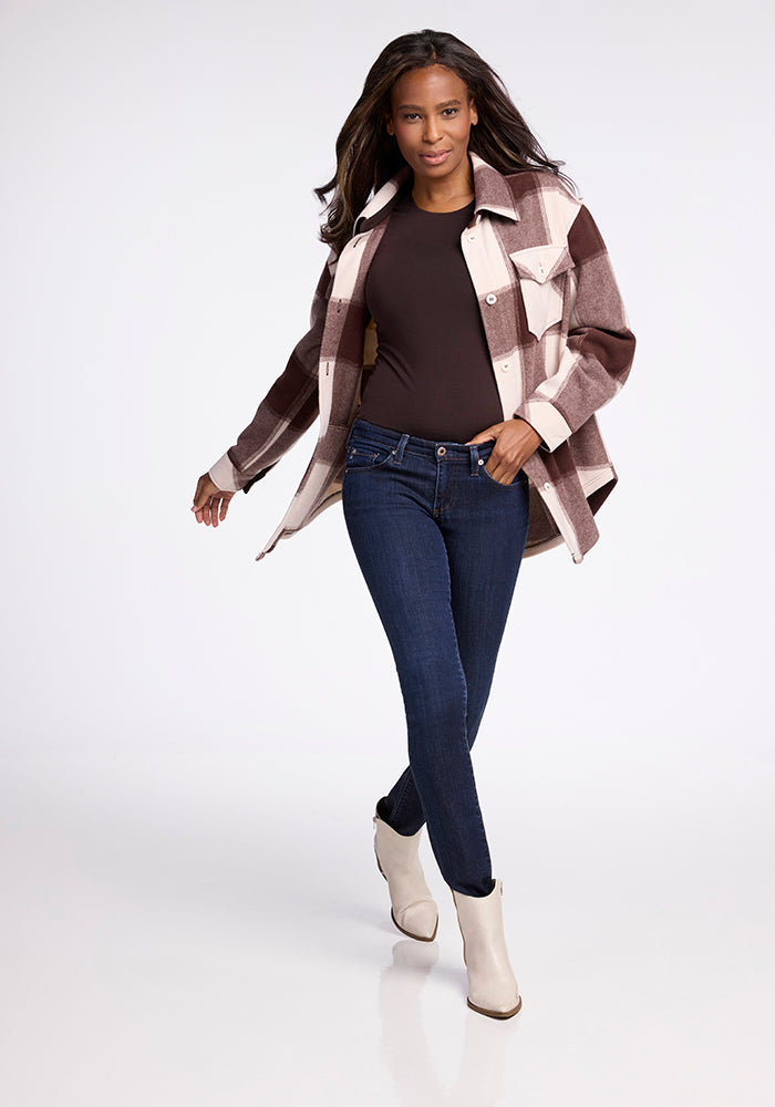A woman with long dark hair models the Woolx Sawyer Shacket in Chai Latte Plaid over a black top, dark blue jeans, and white ankle boots as she walks confidently against a plain white background.