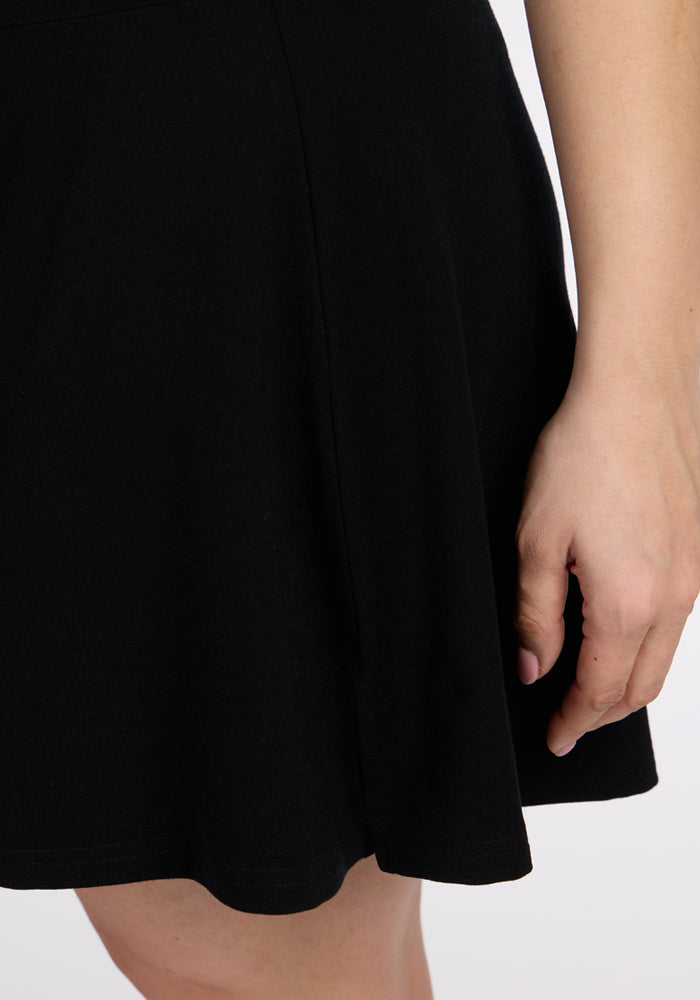 A close-up of a person wearing the Woolx Sienna Skort in black, with their right hand by their side, set against a light background. Only part of the arm and lower torso are visible.