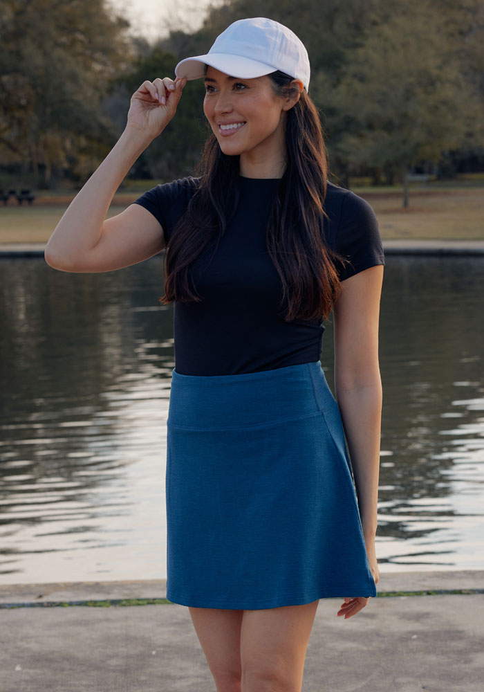 A woman wearing a white cap, black t-shirt, and Woolx Sienna Skort in Majolica Blue smiles by a peaceful lakeside in the park, with trees and grass visible in the background.