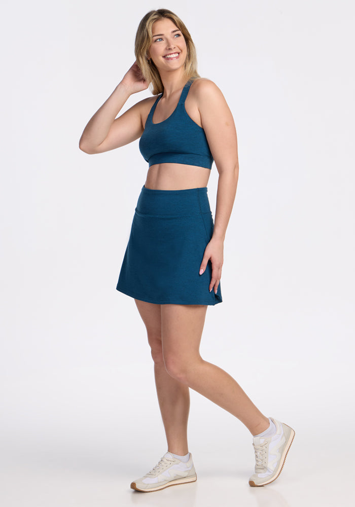 A woman smiles while posing in the Woolx Sienna Skort in Majolica Blue and a teal sports bra, paired with white sneakers, against a plain white background. | Ariana is 5'11", wearing a size S