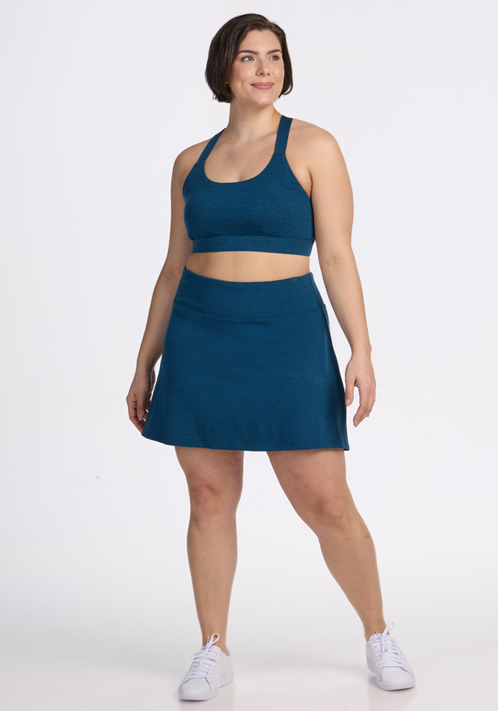 A woman with short brown hair smiles in a Majolica Blue Sienna Skort and matching sports bra by Woolx, paired with white sneakers, against a plain white background. | Juliana is 5'10", wearing a size XL