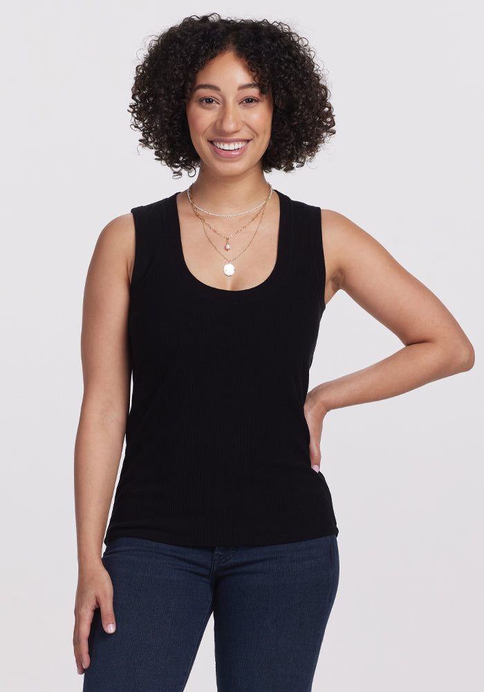 A person with curly hair smiles against a plain background, highlighting the versatile fit of their Sloane Ribbed Tank in black by Woolx. Paired with blue jeans and layered necklaces, it creates a stylish yet effortless look. | Tori is 5'7", wearing a size S