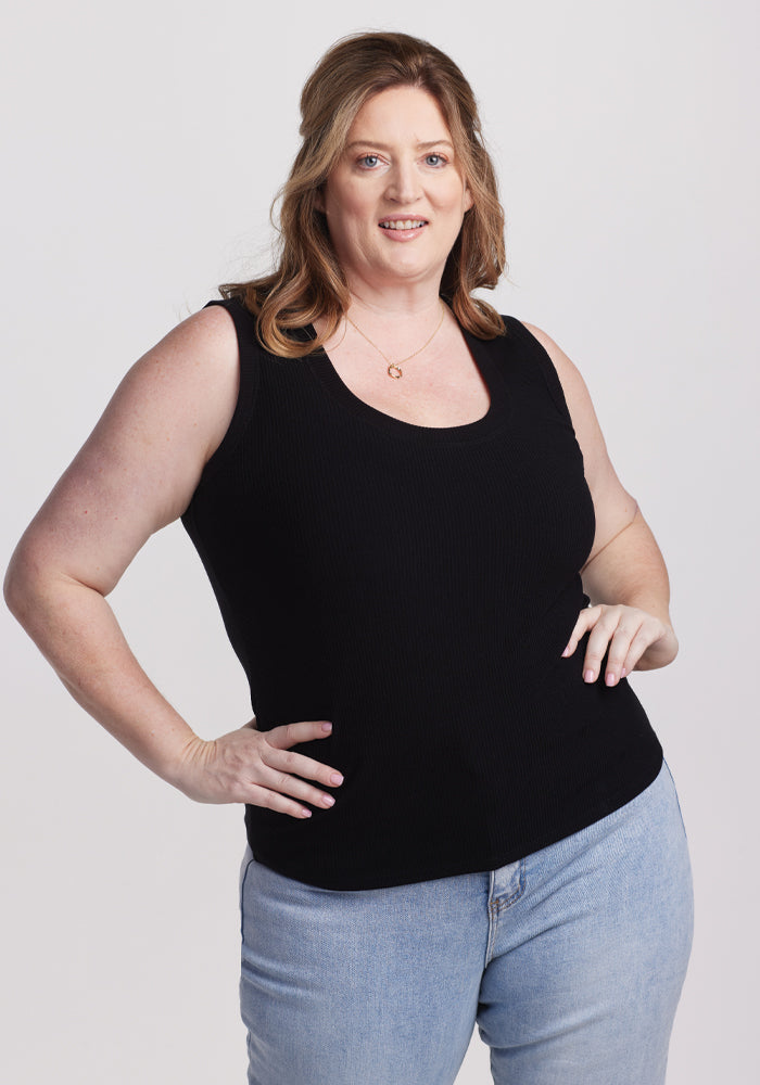 A person with long hair smiles with one hand on their hip against a plain white background, wearing a Woolx Sloane Ribbed Tank in black and light blue jeans. | Cambre is 5'11", wearing a size XL