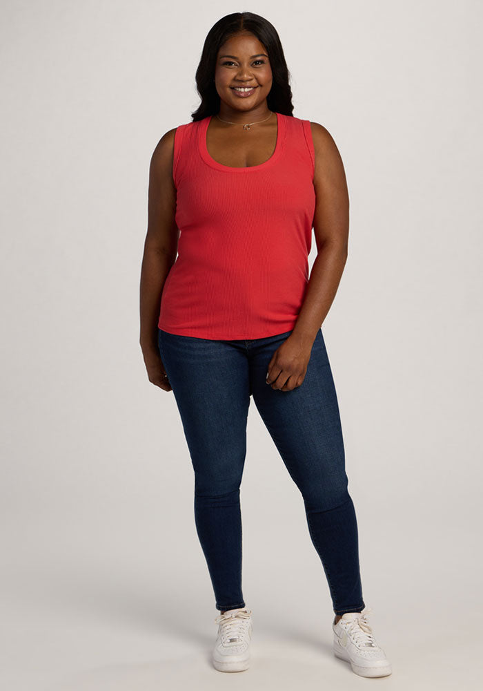 Woman smiling in the Woolx Sloane Ribbed Tank - Hibiscus, blue jeans, and white sneakers against a plain background.