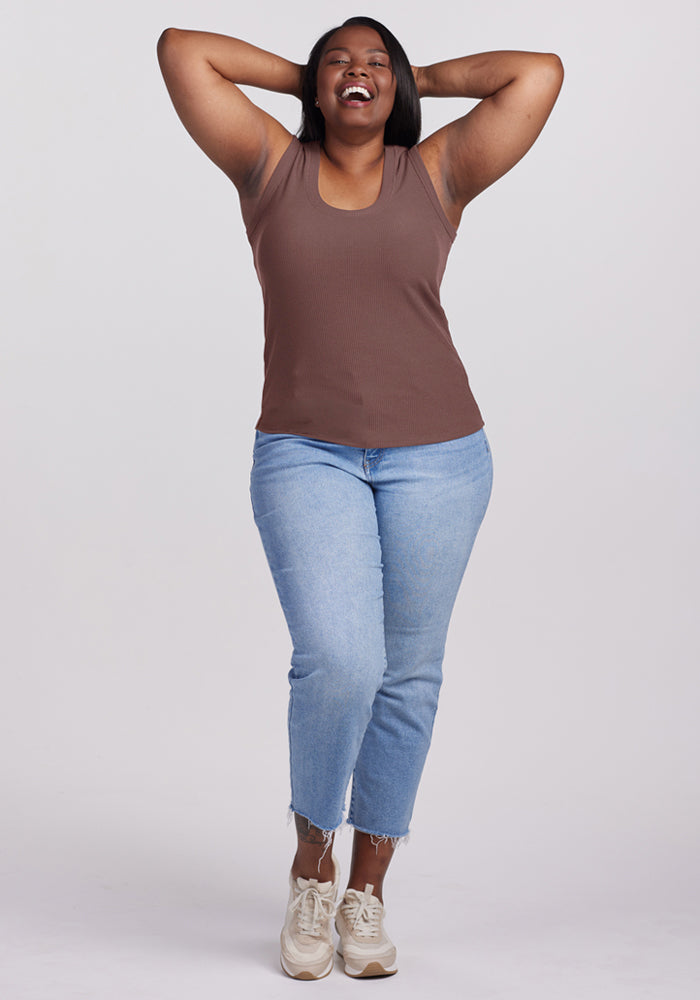 Woman smiles with arms behind her head, wearing a Woolx Sloane Ribbed Tank in Peppercorn, light blue jeans, and beige sneakers.